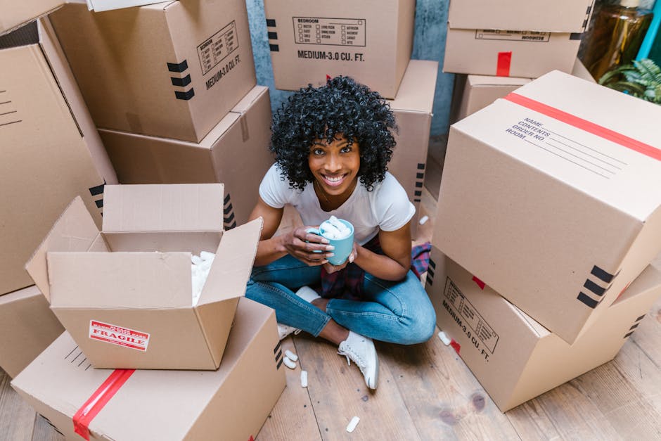 A young woman with curly black hair, smiling while sitting on a wooden floor surrounded by packed cardboard boxes of various sizes, some labeled with contents and fragile markings. She is holding a blue mug with foam or whipped cream inside. The boxes contain packing materials such as white packing paper and plastic wrap, and have red and black printed labels. In the background, more boxes are stacked inside a room, indicating a home packing or moving process as part of a home relocation or furniture transport service. The scene reflects the packing stage of house removals, with natural lighting highlighting the cluttered environment and the woman's cheerful expression during the packing and moving preparations, consistent with removals activities described on manandavanfulham.co.uk.