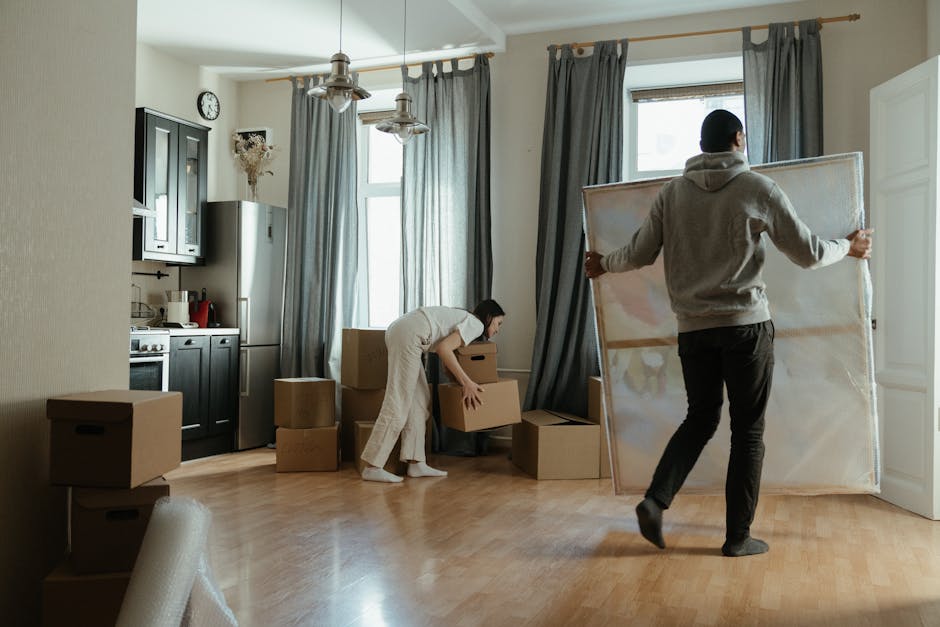 In a well-lit room with a wooden floor, two movers are engaged in an ongoing home relocation process. One individual, dressed in a grey hoodie and black pants, is carrying a large, wrapped piece of furniture, possibly a mirror or framed artwork, towards the right side of the room. The second person, wearing a beige jumpsuit and white socks, is bending down to pick up or position a cardboard box amidst several similar boxes stacked on the floor. The background features a window with natural light filtering through grey curtains, a wall-mounted clock, and kitchen units including a refrigerator, oven, and cupboards. Various packing materials, such as brown cardboard boxes, some partial foam padding, and clear plastic wrap, are visible, indicating an active packing and moving process. [COMPANY_NAME] is involved in this scene, supporting the logistics of furniture transport and packing during the flat move near Fulham Broadway station, aligned with professional house removals and moving services.