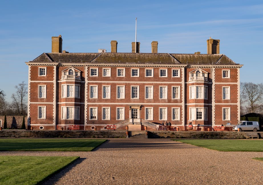 A large, historic brick mansion with multiple windows and decorative white stone accents, situated on a well-maintained lawn with a wide stone staircase leading to the main entrance. The building feat