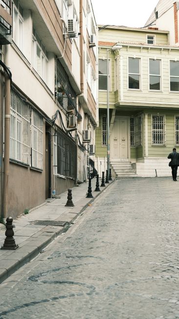 A narrow cobblestone street in Fulham, lined with multi-storey residential buildings featuring modern and traditional architectural elements. On the left, beige and grey building facades display windows with security bars and external air conditioning units mounted on the walls. The right side shows a green, wooden-clad property with bay windows, a staircase leading to the entrance, and a small porch area. A black man with a suit and tie is walking away from the camera towards the end of the street. The street is enclosed with short black bollards along the pavement edge, and the scene is lit with natural daylight. This setting exemplifies urban house relocation challenges, such as narrow passages typical of FULHAM, which [COMPANY_NAME] might navigate when providing removals or furniture transport services for house moves in confined city streets.