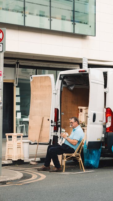 A man sitting on a wooden chair outside a white van during a home relocation process on New Kings Road, Fulham. The van's rear doors are open, revealing wooden furniture pieces, including a large panel leaning against the vehicle, and cardboard boxes inside ready for transport. The man is holding and inspecting a small item, possibly a packing material or document, as part of the packing and moving activities. The scene is set on a paved street with the van parked very close to the building's entrance, which has large glass windows and an upper-story balcony. Visible equipment includes a small wooden crate and packing materials, indicating preparation for furniture transport. The lighting suggests it is daytime, and the environment reflects a typical urban setting for professional removals, supported by [COMPANY_NAME] and its scheduled services for house and furniture removals along the New Kings Road area.