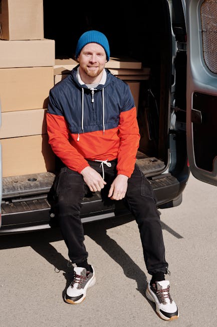 A young man with a beard and fair skin, wearing a blue beanie, a blue and red waterproof jacket, black pants, and white sneakers, is sitting on the open rear door of a black van parked on a street. Inside the van, multiple cardboard boxes of various sizes are visible, some wrapped in plastic or fabric for protection during home relocation. The boxes are stacked against the van's interior walls, which are dark, and some boxes are placed on a wooden pallet or base for easier handling. The man is smiling and appears relaxed, possibly taking a break during the furniture transport or packing and moving process. The van is positioned in a well-lit outdoor setting, with a paved surface underneath. This scene exemplifies a professional moving service such as that provided by manandavanfulham.co.uk, involving loading or unloading boxes as part of house removals on the New Kings Road in Fulham.