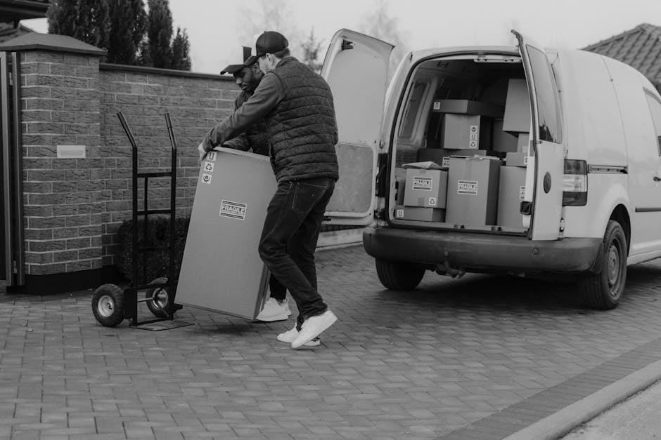 A man sitting on a wooden chair outside a white van during a home relocation process on New Kings Road, Fulham. The van's rear doors are open, revealing wooden furniture pieces, including a large panel leaning against the vehicle, and cardboard boxes inside ready for transport. The man is holding and inspecting a small item, possibly a packing material or document, as part of the packing and moving activities. The scene is set on a paved street with the van parked very close to the building's entrance, which has large glass windows and an upper-story balcony. Visible equipment includes a small wooden crate and packing materials, indicating preparation for furniture transport. The lighting suggests it is daytime, and the environment reflects a typical urban setting for professional removals, supported by [COMPANY_NAME] and its scheduled services for house and furniture removals along the New Kings Road area.